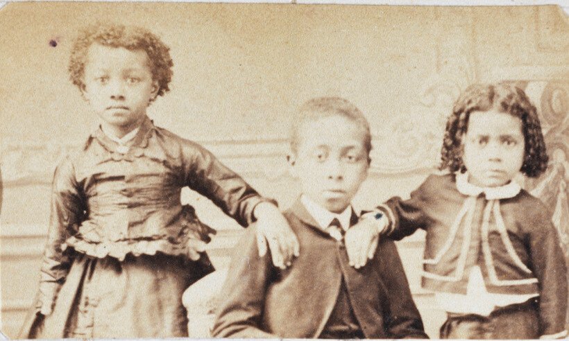 Fotografia do século XIX, em tom sépia, feita por Militão Augusto de Azevedo. A imagem mostra três crianças negras posando diante de um fundo neutro com decoração clássica. À esquerda, uma menina está em pé, com a mão apoiada no ombro do menino ao centro; ela veste um vestido escuro com babados e tem cabelo crespo curto. No centro, sentado, está um menino vestindo paletó, colete e gravata, com expressão séria. À direita, outra menina, também com expressão séria, está sentada com o braço apoiado no ombro do menino; ela tem cabelo encaracolado e veste um conjunto escuro com gola branca e laço. Abaixo da imagem, há a inscrição manuscrita '4456'. A fotografia faz parte do acervo do Museu Paulista da Universidade de São Paulo e documenta com sensibilidade a presença da infância negra na sociedade brasileira oitocentista.