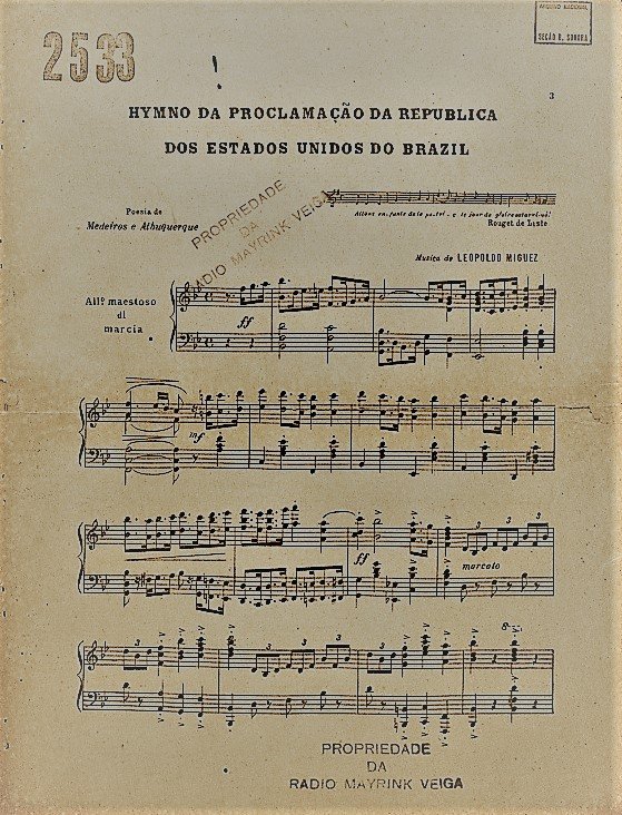 Partitura impressa do final do século XIX com o título 'Hymno da Proclamação da República dos Estados Unidos do Brazil', com grafia da época. No topo, há o número 2533 em destaque. Abaixo do título, os créditos indicam a poesia de Medeiros e Albuquerque e a música de Leopoldo Miguez, então diretor do Instituto Nacional de Música. A partitura contém cinco linhas com pautas e notas musicais. Há anotações de andamento como 'All° maestoso di marcia'. Ao centro e na base da página aparece a marca 'Propriedade da Rádio Mayrink Veiga' em carimbo roxo. O papel amarelado apresenta marcas do tempo, reforçando sua antiguidade.