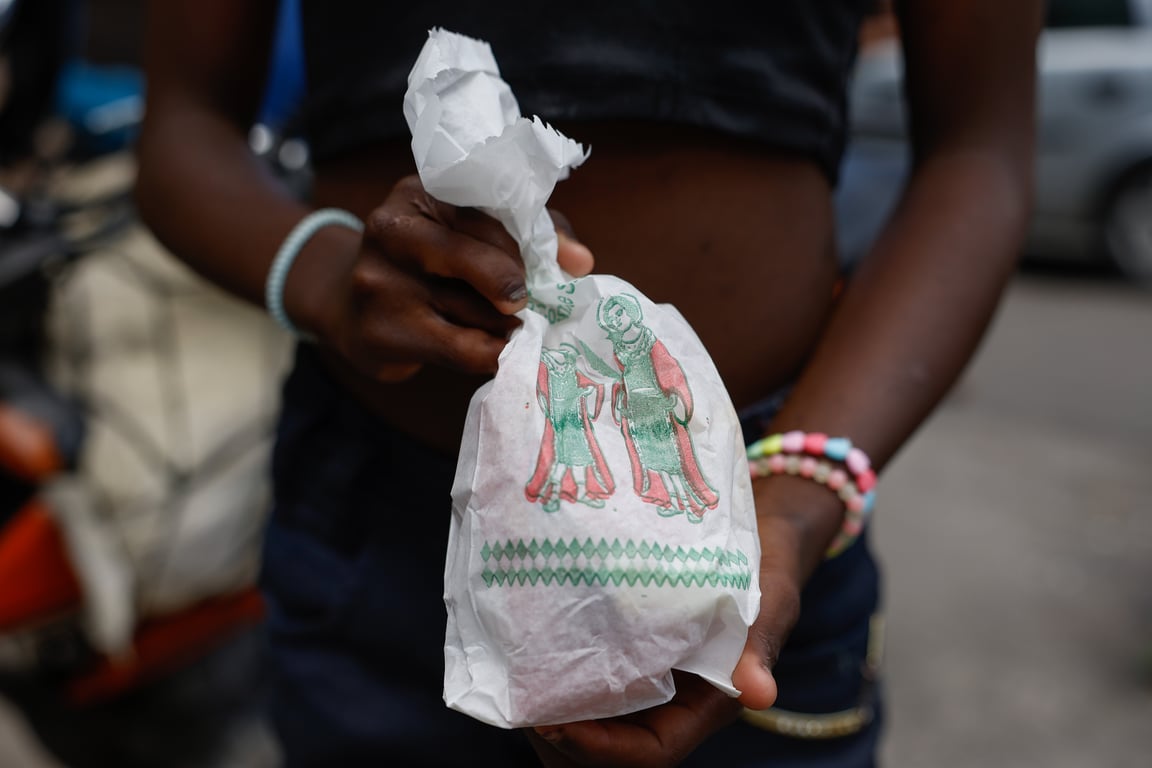 Fotografia do Dia de Cosme e Damião, de Fernando Frazão, produzida para a Agência Brasil. A imagem mostra, em primeiro plano, as mãos de uma pessoa negra segurando um pequeno pacote embrulhado em papel branco. Na embalagem há uma ilustração colorida dos santos Cosme e Damião. A pessoa veste roupa escura e usa pulseiras coloridas nos pulsos. O enquadramento destaca apenas parte do corpo, sem mostrar o rosto. Ao fundo, a cena aparece desfocada, revelando uma rua com veículos e pessoas. A fotografia registra a tradicional distribuição de doces realizada nas celebrações em homenagem a Cosme e Damião.