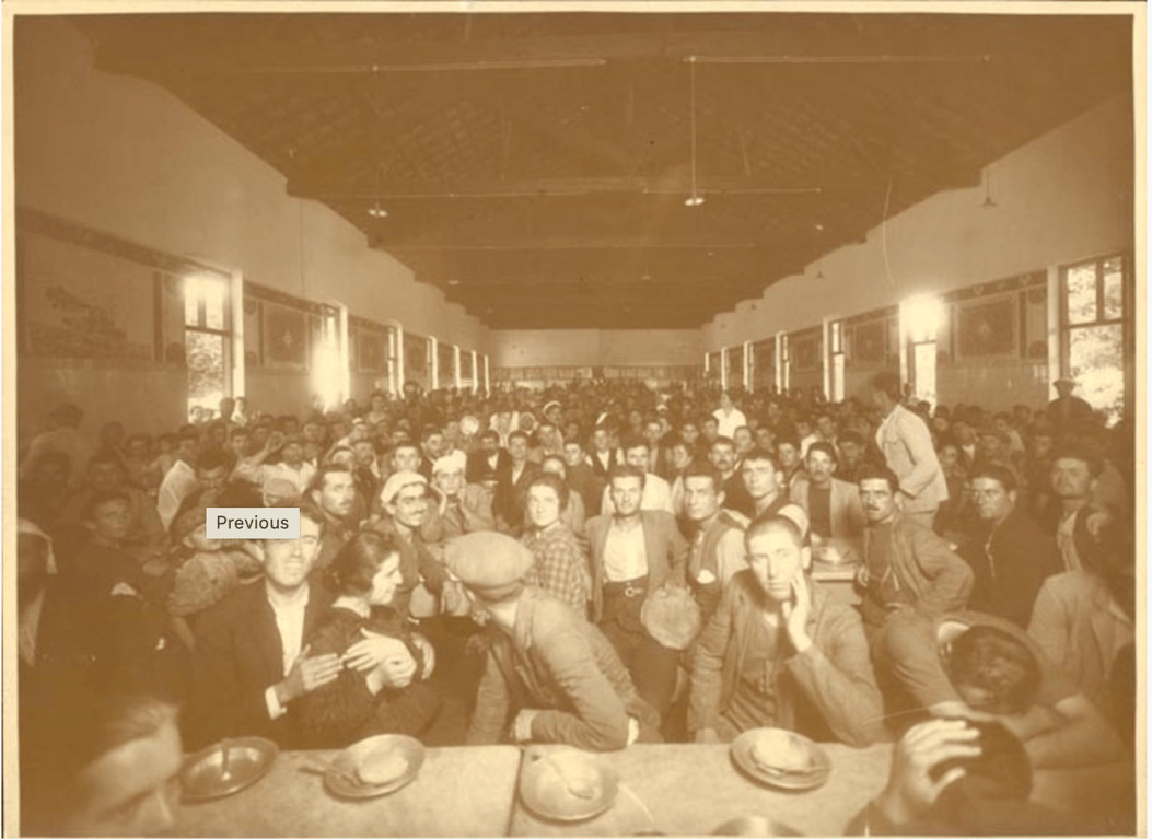 Fotografia em tom sépia, tirada no interior de um grande refeitório com pé-direito alto e teto de madeira. A imagem mostra um salão lotado de pessoas sentadas lado a lado em longas mesas coletivas. A maioria são homens, com roupas simples — muitos usando bonés ou boinas —, e alguns poucos rostos femininos são visíveis à frente. Os olhares estão voltados diretamente para a câmera, e as expressões variam entre cansaço, desconfiança e curiosidade. À frente, sobre a mesa, há pratos e tigelas vazias. O ambiente tem janelas nas duas laterais, por onde entra luz natural. Trata-se do refeitório da Hospedaria de Imigrantes na Ilha das Flores, no Rio de Janeiro — um espaço de acolhimento temporário para estrangeiros recém-chegados ao Brasil, em funcionamento especialmente entre os séculos XIX e XX. Fotografia de autor desconhecido, pertencente à Coleção Leopoldino Brasil.