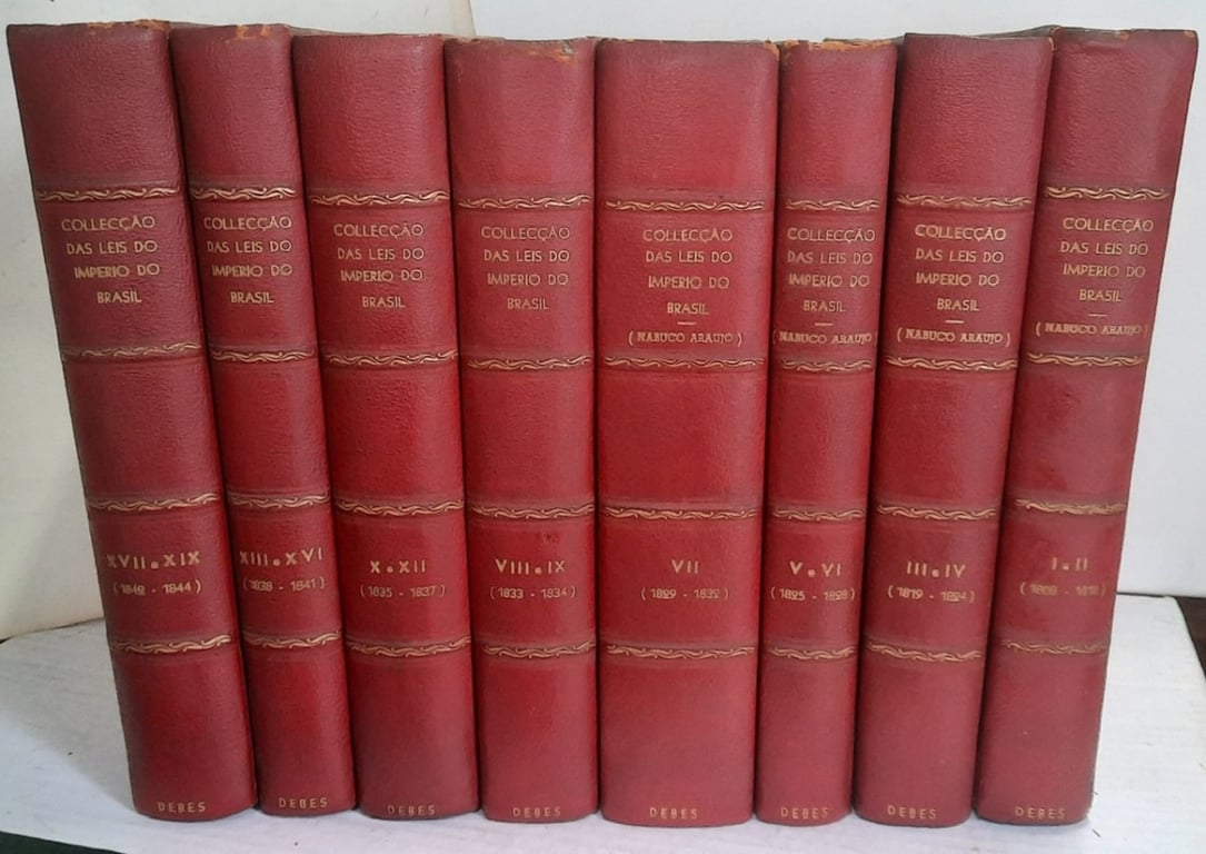 A imagem mostra oito livros antigos, encadernados em couro vermelho, alinhados lado a lado. As lombadas são decoradas com detalhes dourados e trazem a inscrição “Collecção das Leis do Império do Brasil”. Os volumes estão organizados por diferentes períodos de anos, abrangendo de 1808 a 1844. Em alguns deles aparece também o nome “Nabuco Araújo”. Os livros estão apoiados sobre uma superfície clara, diante de um fundo branco.