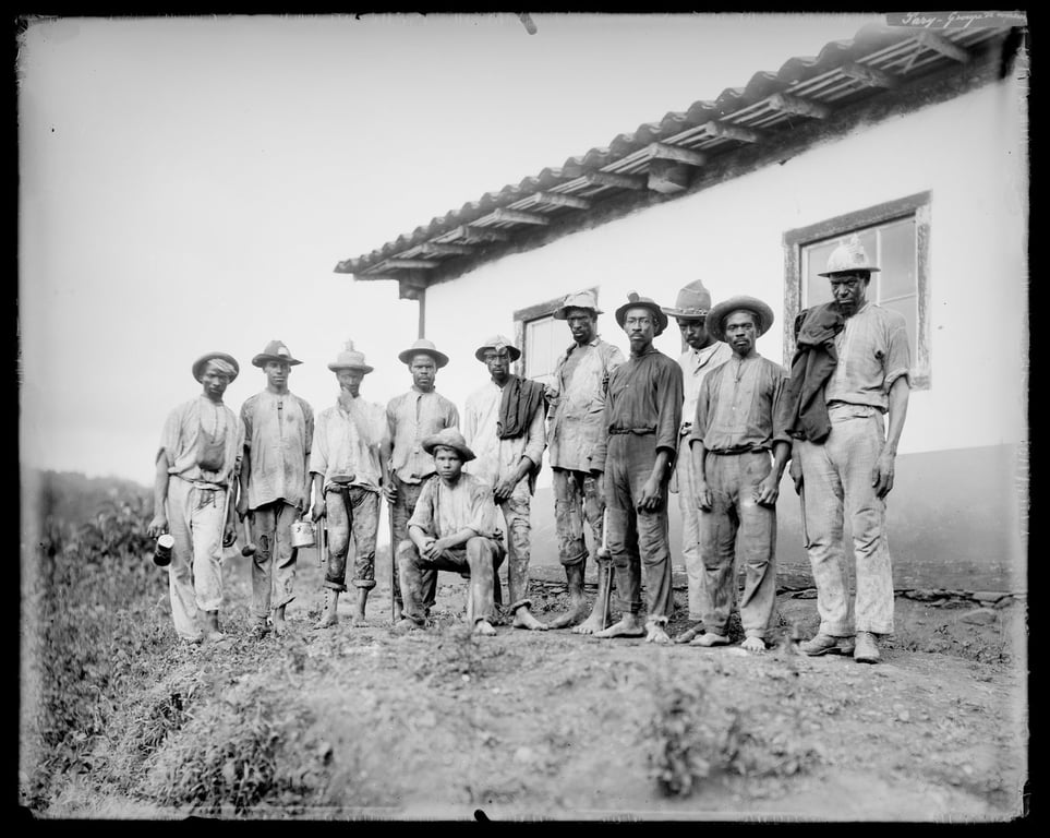 fotografia em preto e branco de um grupo de mineiros, registrada em 1885 por Marc Ferrez, em Minas Gerais, Brasil, que captura o contexto do trabalho exaustivo na mineração durante o século XIX. O grupo, composto por homens negros, veste roupas simples e gastas, com calças e camisas de mangas compridas, algumas arregaçadas. Muitos usam chapéus de palha ou de aba larga. A maioria está de pé, com expressões sérias, enquanto um homem se encontra agachado em frente ao grupo. O cenário é rústico, com chão de terra batida e um barracão ao fundo, com paredes brancas e telhado de barro.