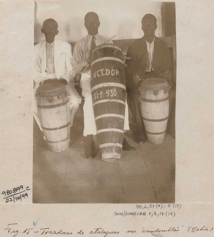 Fotografia em preto e branco da Coleção Melville Herskovits, tirada na Bahia entre as décadas de 40 e 60. A imagem mostra três homens negros posicionados diante de uma parede simples, em um ambiente fechado, sentados lado a lado, cada um com um atabaque entre as pernas. Eles usam roupas sociais, dois deles com paletó e o do centro com camisa listrada clara. A fotografia possui anotações manuscritas na borda inferior e lateral. No rodapé, em letra cursiva, lê-se: 'Tocadores de atabaques no candomblé (Bahia)'.