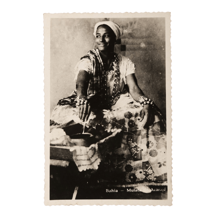 Fotografia em preto e branco mostra uma mulher negra sorridente, sentada ao lado de um tabuleiro com comidas típicas. Ela veste uma tradicional roupa de baiana: saia rodada com estampa floral, blusa rendada, pano da costa, colares e pulseiras, além de um turbante branco na cabeça. A mulher representa a figura histórica da vendedora ambulante de comidas afro-baianas, símbolo da resistência cultural e religiosa do povo negro no Brasil.