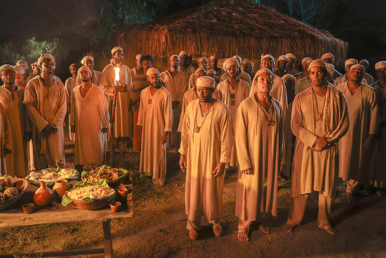 A imagem é uma cena do filme Malês, de 2024, dirigido por Antonio Pitanga, que retrata a maior insurreição de escravizados da história do país: a Revolta dos Malês. Trata-se de uma fotografia noturna, com iluminação quente de tochas, mostrando um grande grupo de pessoas negras reunidas em frente a uma cabana de palha. A maioria veste roupas longas e claras, em tons de bege e branco, e algumas pessoas usam turbantes ou lenços na cabeça. No centro e à frente estão três homens em destaque, olhando para a câmera. À esquerda, há uma mesa de madeira com diversos alimentos, frutas, folhas e recipientes. Ao fundo, a cabana tem paredes e cobertura feitas de palha, cercada por vegetação.