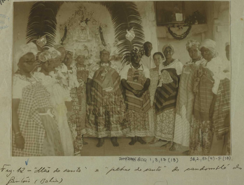Fotografia registrada em Salvador (Bahia), em 1940, por Arthur Ramos. Mostra um grupo de mulheres e homens negros reunidos em frente a um altar religioso, decorado com imagens e elementos simbólicos das religiões de matriz africana. Ao centro da imagem está a “Mãe do santo” - estrutura ornamentada com penas e elementos sagrados, atrás de um conjunto de pessoas com trajes tradicionais do candomblé, como saias rodadas, panos da costa, colares e turbantes. Algumas das mulheres estão de mãos dadas ou lado a lado, formando uma fileira. A imagem representa o contexto das práticas religiosas afro-brasileiras, intensamente perseguidas no início do século XX.
