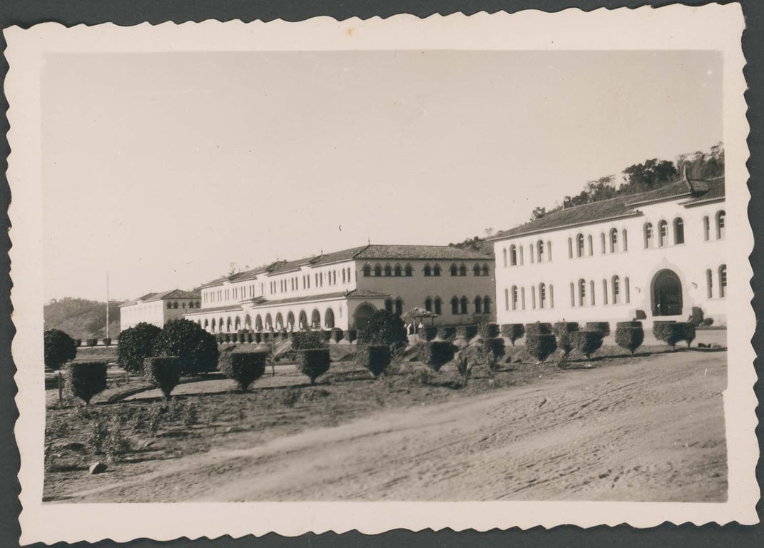 Fotografia em preto e branco, com bordas onduladas, típica de registros do início a meados do século XX. A imagem mostra um grande edifício institucional de dois andares, com fachada branca, janelas em arco e telhado de telhas cerâmicas. À esquerda, uma longa galeria com colunatas marca a entrada principal do complexo. A construção se estende horizontalmente, cercada por um jardim com árvores e arbustos simetricamente podados. O terreno em frente ao prédio é de terra batida, indicando que a área ainda estava em desenvolvimento urbano. Ao fundo, vê-se um morro com vegetação nativa. Trata-se de um patronato agrícola, instituição criada para abrigar, educar e disciplinar jovens, especialmente órfãos ou considerados 'menores em situação irregular', sob o pretexto de regeneração por meio do trabalho no campo – prática comum nas políticas assistencialistas e repressivas do Brasil do século XX.