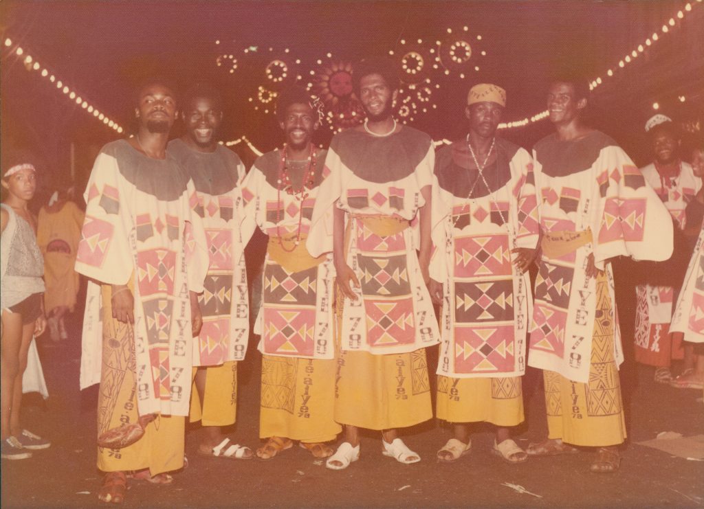 Fotografia colorida, provavelmente dos anos 1970 ou início dos anos 1980, mostrando o desfile do bloco afro Ilê Aiyê durante o carnaval de Salvador. A imagem captura dezenas de integrantes do bloco, todos negros, vestindo trajes tradicionais africanos em tons vibrantes de vermelho, amarelo e branco, com estampas geométricas e turbantes elaborados. Os foliões estão em movimento na rua, alguns tocando instrumentos de percussão, outros dançando e cantando. Ao fundo, vê-se uma multidão de espectadores observando o desfile. A atmosfera é de celebração, orgulho negro e afirmação da identidade afro-brasileira durante um período de intensa repressão da ditadura militar.