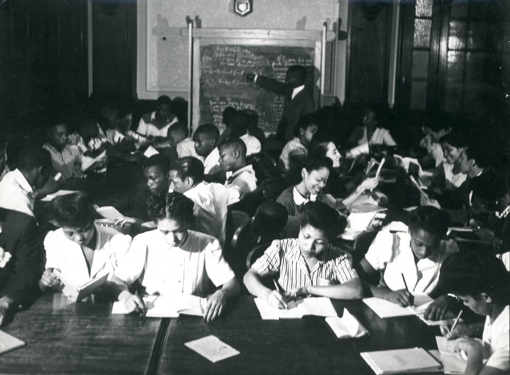 Fotografia em preto e branco da sala de aula do TEN - Teatro Experimental do Negro, repleta de estudantes negros, sentados lado a lado em longas mesas de madeira, com cadernos e livros abertos à sua frente. Na parte central da imagem, várias mulheres escrevem com atenção, enquanto outras pessoas leem ou observam seus materiais de estudo. Ao fundo, o professor Ironides Rodrigues, homem negro em pé, de terno escuro, aponta para o quadro-negro repleto de anotações escritas à mão. O ambiente é de concentração e aprendizado coletivo, transmitindo a importância da educação como ferramenta de emancipação.