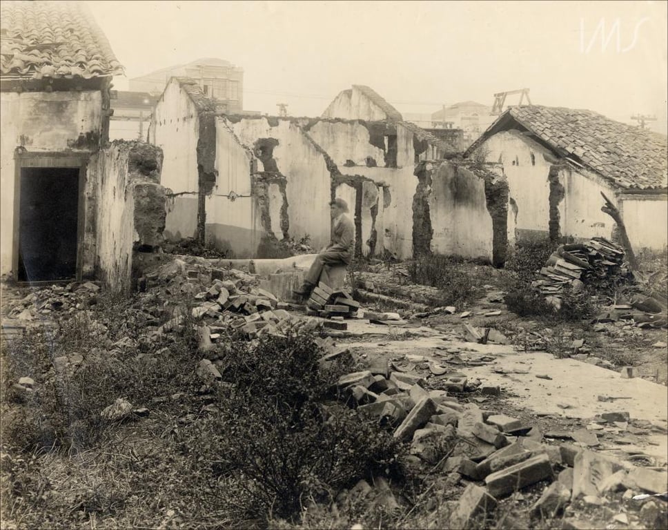 Fotografia em preto e branco de Vincenzo Pastore, acervo Instituto Moreira Salles. de um conjunto de casas em ruínas, registradas durante processo de demolição. As paredes estão parcialmente destruídas, com vãos abertos onde antes havia portas e janelas. A maior parte dos telhados está ausente ou desabada, restando apenas a estrutura de algumas casas. No centro da imagem, um homem branco sentado em um sofá entre os escombros, com expressão neutra. O chão está coberto por tijolos, pedras e mato rasteiro. Ao fundo, vê-se a silhueta de prédios desfocados, sugerindo que a foto foi tirada em área urbana.