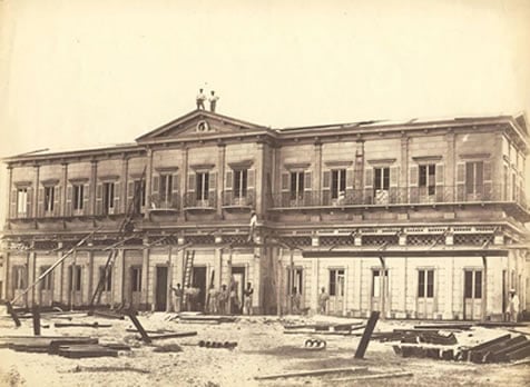 Fotografia histórica da fachada da Estação de Jequitaia, na Bahia, pertencente à Coleção Vignoles da Instituição de Engenheiros Civis de Londres. A imagem mostra um prédio comprido de dois andares, com fachada simétrica e diversas portas e janelas distribuídas ao longo de toda a construção. No centro da parte superior, há um frontão triangular com um pequeno ornamento no topo. Em frente ao edifício, o terreno é amplo e sem pavimentação, com trilhos, vigas de madeira e outros materiais espalhados, indicando obras ou a implantação da ferrovia. Pequenos grupos de pessoas aparecem próximos à entrada principal, enquanto a fotografia, em tons de sépia, registra um dos primeiros conjuntos ferroviários construídos na Bahia.