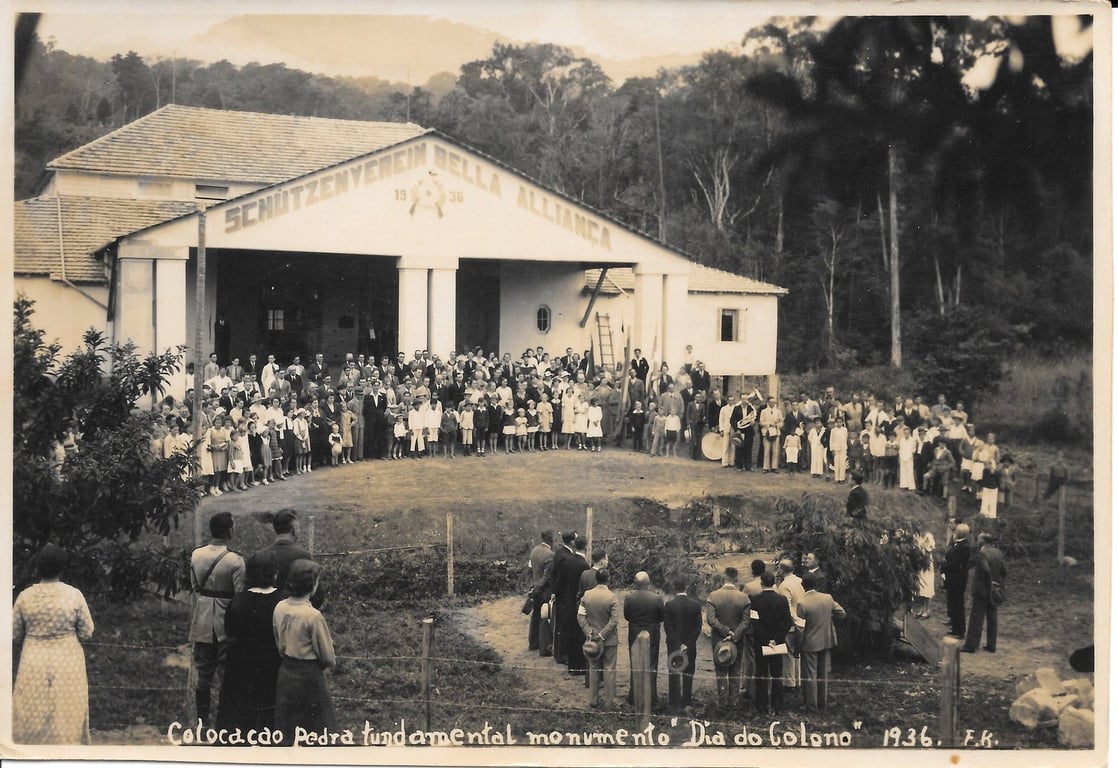 Fotografia em preto e branco registra uma celebração no Clube de Caça e Tiro, em Rio do Sul, Santa Catarina, no ano de 1936. Um grupo de pessoas, provavelmente imigrantes alemães, posa para a foto ao ar livre. Homens usam trajes sociais, alguns com suspensórios e chapéus, enquanto mulheres vestem vestidos longos e claros. Crianças também estão presentes na imagem. Ao fundo, há uma grande faixa pintada na parede com 'schutzenverein bela aliança', que são, em países germânicos, associações de tiro.