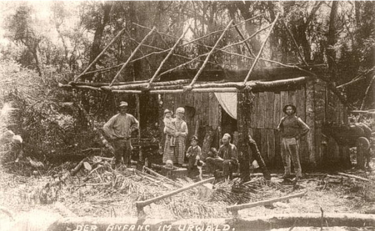 Fotografia em sépia de uma família de colonos alemães posando diante de uma casa de madeira em meio à mata. Em primeiro plano, troncos de árvore no chão e ferramentas de trabalho rural sugerem que a casa está em construção, com a estrutura do telhado ainda incompleta. Algumas pessoas estão em frente à casa: homens, mulheres e crianças, todas vestindo roupas típicas do período. Um homem está de pé com as mãos na cintura, outro segura uma criança no colo. A imagem expressa o processo de ocupação do território e construção de moradias.