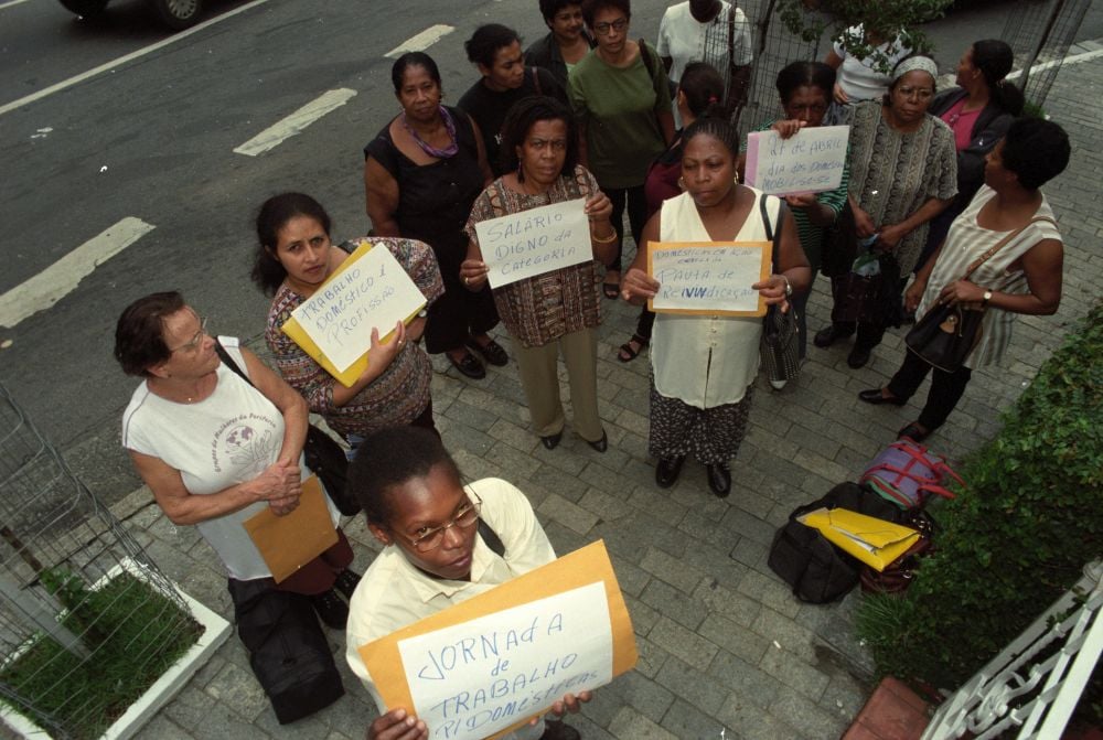 A fotografia aérea mostra um grupo de mulheres em protesto, segurando cartazes com frases que se relacionam a direitos trabalhistas e salariais para empregadas domésticas, com frases como 'Salário digno na categoria' e 'Jornada de trabalho p/ domésticas'.
