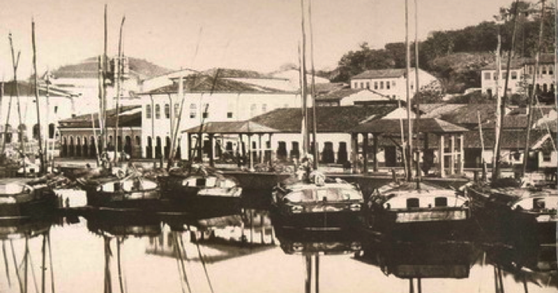 Fotografia histórica do Porto da Vila de Nazaré das Farinhas, de 1865, registrada por Camillo Vedani. A imagem mostra um conjunto de embarcações de madeira ancoradas às margens do porto, com mastros altos refletidos na água calma. Ao fundo, aparecem armazéns, casarões e outras construções voltadas para a orla, formando o núcleo urbano da vila. Mais acima, sobre a encosta, há outras edificações cercadas por árvores e vegetação. A fotografia, em tons de sépia, registra o porto de Nazaré das Farinhas, um importante ponto de circulação de pessoas e mercadorias no Recôncavo Baiano durante o século XIX.