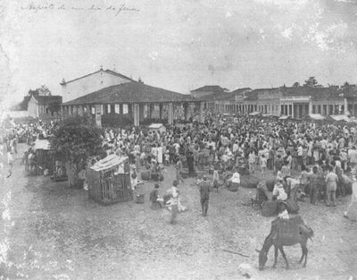 Fotografia em preto e branco da feira livre do município de Santo Antônio de Jesus, registrada no início do século XX e pertencente ao Arquivo da Família Tourinho. A imagem mostra uma grande praça ocupada por centenas de pessoas reunidas em dia de feira. No centro, destaca-se um amplo mercado coberto, cercado por barracas, carroças e vendedores. Em diferentes pontos da praça, homens, mulheres e crianças circulam, conversam ou permanecem próximos aos produtos expostos. No primeiro plano, à direita, um homem conduz um animal de carga carregado com mercadorias. Ao fundo, aparecem construções térreas e sobrados que contornam a praça. A fotografia registra a intensa movimentação comercial e o papel da feira livre como importante espaço de encontro e abastecimento da população de Santo Antônio de Jesus.