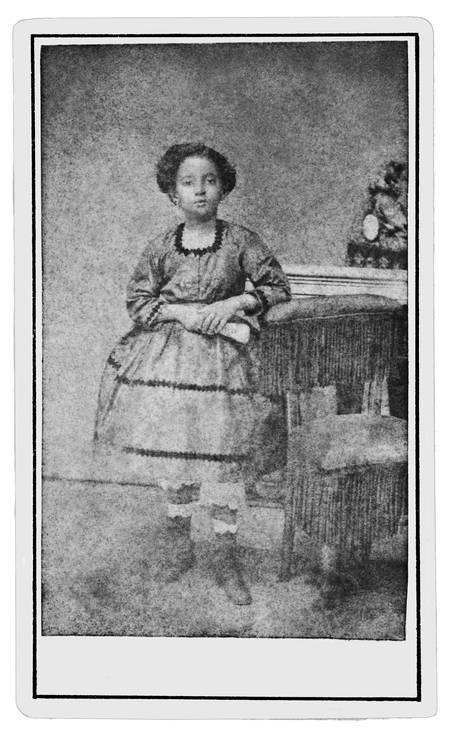 Fotografia em preto e branco, com moldura branca, mostrando Amália Augusta, mãe do escritor Lima Barreto. Ela é uma mulher negra, de pele clara, aparentando ser jovem, com cabelos curtos e cacheados. Está em pé, levemente voltada para a direita, ao lado de uma mesinha coberta por um tecido escuro com franjas. Usa vestido de mangas longas e saia rodada, com detalhes em faixas horizontais escuras e gola rendada. Ela segura um pequeno objeto claro, possivelmente um lenço ou livro, entre as mãos cruzadas à frente do corpo. Ao fundo, um arranjo de flores decora a mesa. A fotografia tem aparência envelhecida, com aspecto esmaecido e fundo neutro.