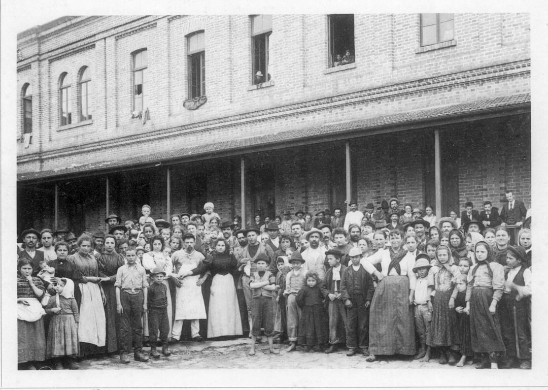 Fotografia em preto e branco de 1890, retratando um grande grupo de imigrantes italianos posando no pátio central da Hospedaria dos Imigrantes, em São Paulo. Homens, mulheres e crianças estão reunidos diante de um prédio de tijolos, com uma varanda no segundo andar e janelas abertas. Alguns usam aventais e roupas de trabalho, enquanto outros vestem trajes mais formais. A fotografia tem um enquadramento amplo, capturando a multidão e a arquitetura da época, registrando o início do fluxo massivo de imigração europeia ao Brasil no final do século XIX.