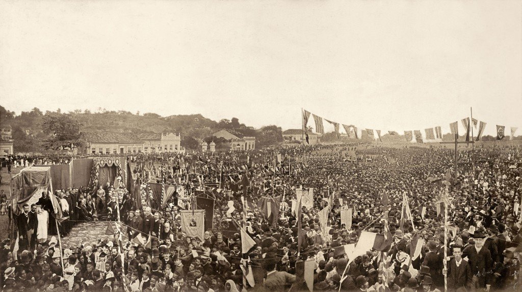 Em uma fotografia histórica em preto e branco, uma multidão enorme se reúne ao ar livre em uma missa campal em 1888, em ação de graças pela Abolição da Escravatura no Brasil. O local está repleto de pessoas de diferentes idades e trajes da época, formando uma densa massa de indivíduos que preenche toda a área. Bandeiras e estandartes são erguidos, adicionando um aspecto de celebração e solenidade ao evento. No centro da imagem, uma estrutura coberta abriga o altar onde os celebrantes conduzem a missa. Ao fundo, pode-se ver edificações e árvores, que delimitam o espaço e dão profundidade à cena. O céu claro e sem nuvens sugere que a missa ocorre durante o dia.