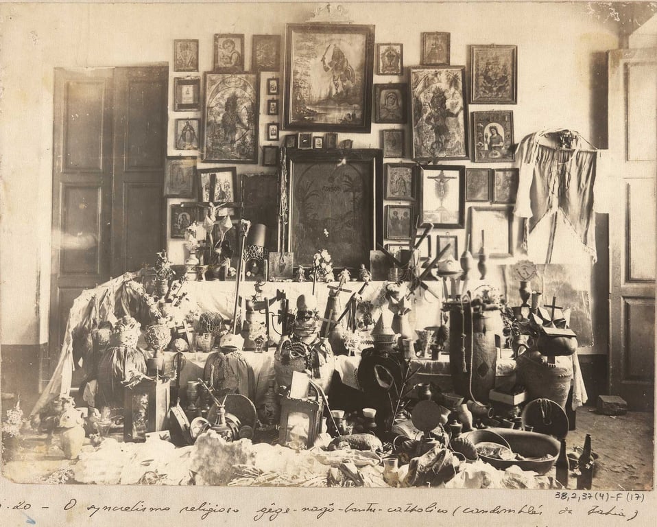 Fotografia em preto e branco tirada por Arthur Ramos em 1940, de um ambiente interno densamente decorado com objetos de culto religioso afro-brasileiro. No centro da imagem, uma grande mesa está coberta por tecido branco e repleta de objetos simbólicos: vasos, imagens, instrumentos, flores, velas e outros elementos ligados a rituais. Ao redor e no chão, há mais objetos, muitos deles grandes e variados, sugerindo diversidade de práticas. Na parede ao fundo, dezenas de quadros estão organizados de forma densa, com imagens religiosas e retratos.