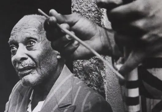 Retrato em preto e branco de Mestre Pastinha, importante referência da capoeira angola. Ele está sentado de perfil, olhando para cima, com expressão serena. Veste um paletó listrado e, à frente, outra pessoa em suas mãos seguram um berimbau. A composição destaca a dignidade e ancestralidade do mestre baiano, que consolidou a capoeira como expressão cultural e pedagógica afro-brasileira.