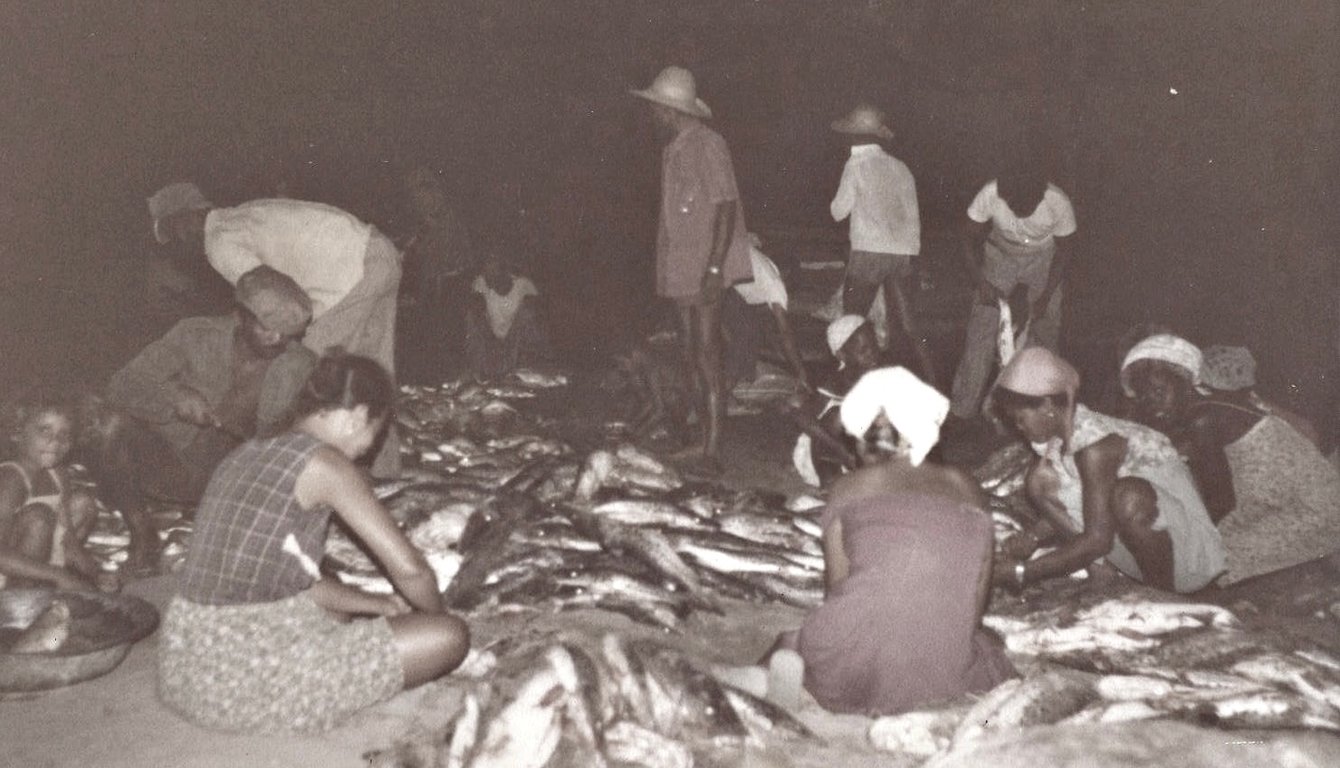 Fotografia histórica da pescaria em Xique-Xique, Bahia, registrada em 1979 e publicada no Blog do Juarez Chaves. A imagem mostra um grupo de homens, mulheres e crianças reunidos durante a separação e limpeza de peixes. Em primeiro plano, mulheres sentadas no chão organizam os peixes espalhados sobre a areia, enquanto uma criança observa a atividade ao lado de uma bacia. Ao centro e ao fundo, homens e mulheres trabalham em pé ou agachados, recolhendo, selecionando e transportando o pescado. Alguns usam chapéus de palha e lenços na cabeça. A fotografia, em tons de preto e branco, registra o trabalho coletivo ligado à pesca artesanal às margens do rio São Francisco.