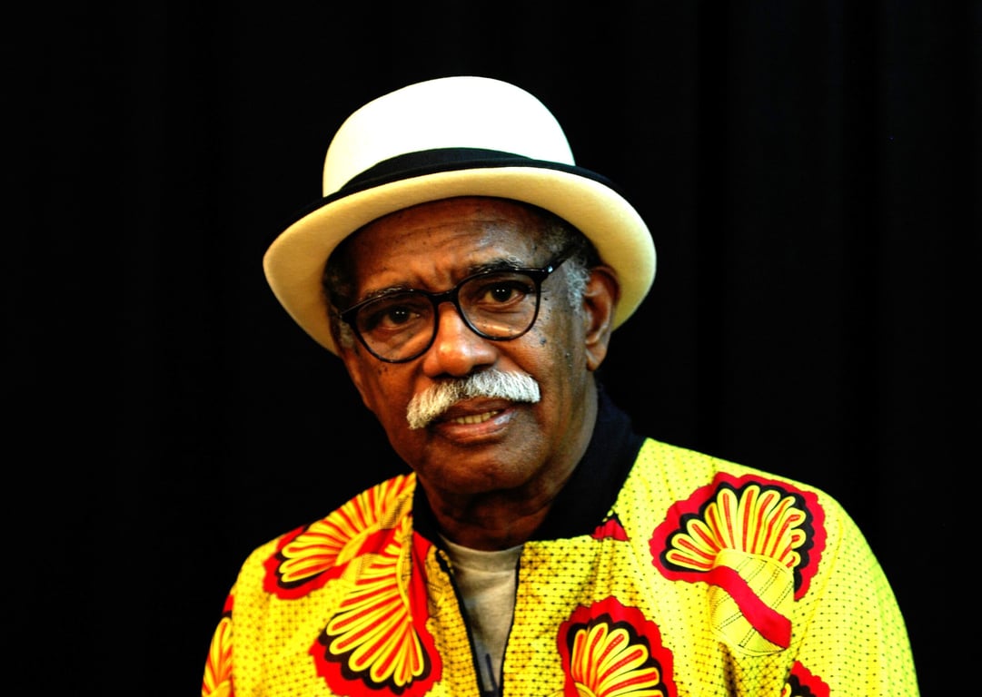 Fotografia de Filó Filho, homem idoso negro, com cabelos grisalhos e bigode, vestindo uma camisa estampada com motivos africanos em tons terrosos. Ele está posicionado em frente a uma parede com grafites coloridos, olhando diretamente para a câmera com uma expressão serena e digna. A imagem transmite a sabedoria e a força de quem viveu décadas de luta pela valorização da cultura negra no Brasil. Ao fundo, é possível ver elementos de arte urbana que remetem à resistência cultural afro-brasileira.