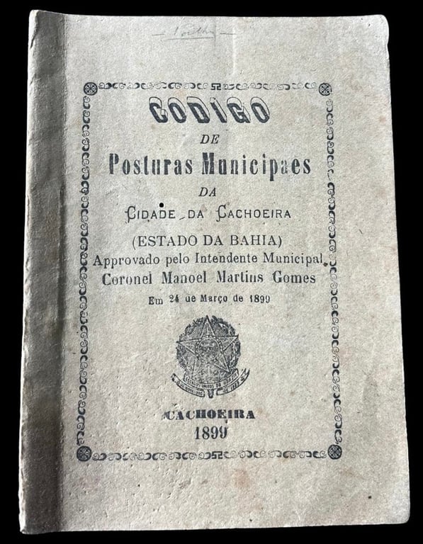 Capa do Código de Posturas Municipais da Cidade da Cachoeira, de 1899, pertencente ao Arquivo Público Municipal de Cachoeira. O documento apresenta uma capa em papel de tom claro, com sinais de envelhecimento e pequenas marcas do tempo. No centro, o título “Código de Posturas Municipais da Cidade da Cachoeira (Estado da Bahia)” aparece em letras pretas de diferentes tamanhos. Logo abaixo, está a informação de que o documento foi aprovado pelo intendente municipal Coronel Manoel Martins Gomes, em 24 de março de 1899. Na parte inferior, aparece o brasão da República e, abaixo dele, a inscrição “Cachoeira 1899”. O texto é contornado por uma moldura decorativa impressa em preto, que acompanha as bordas da capa.