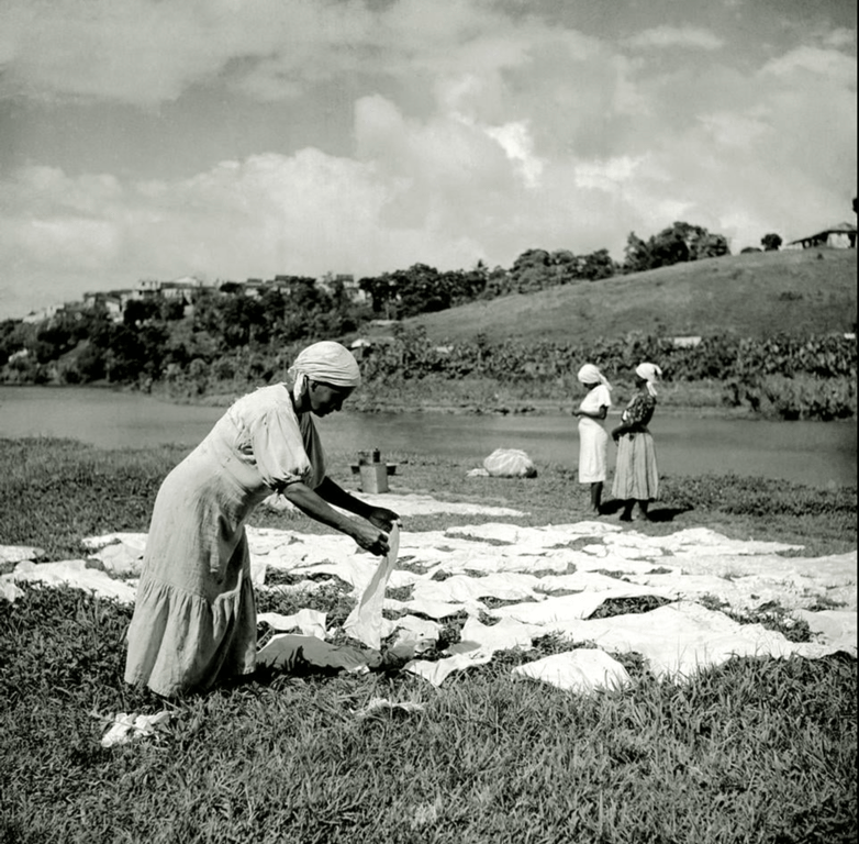 Fotografia em preto e branco de três mulheres negras trabalhando às margens de um rio ou lago, em uma área de campo aberto. No primeiro plano, uma delas, vestida com um vestido claro e turbante, estende ou recolhe roupas brancas dispostas sobre a grama para secar. Ao fundo, outras duas mulheres, também de saias e turbantes, estão em pé conversando, próximas à água. A paisagem mostra colinas verdes, árvores e algumas casas no horizonte. A cena retrata o trabalho doméstico de lavadeiras, atividade historicamente associada às mulheres negras no Brasil.