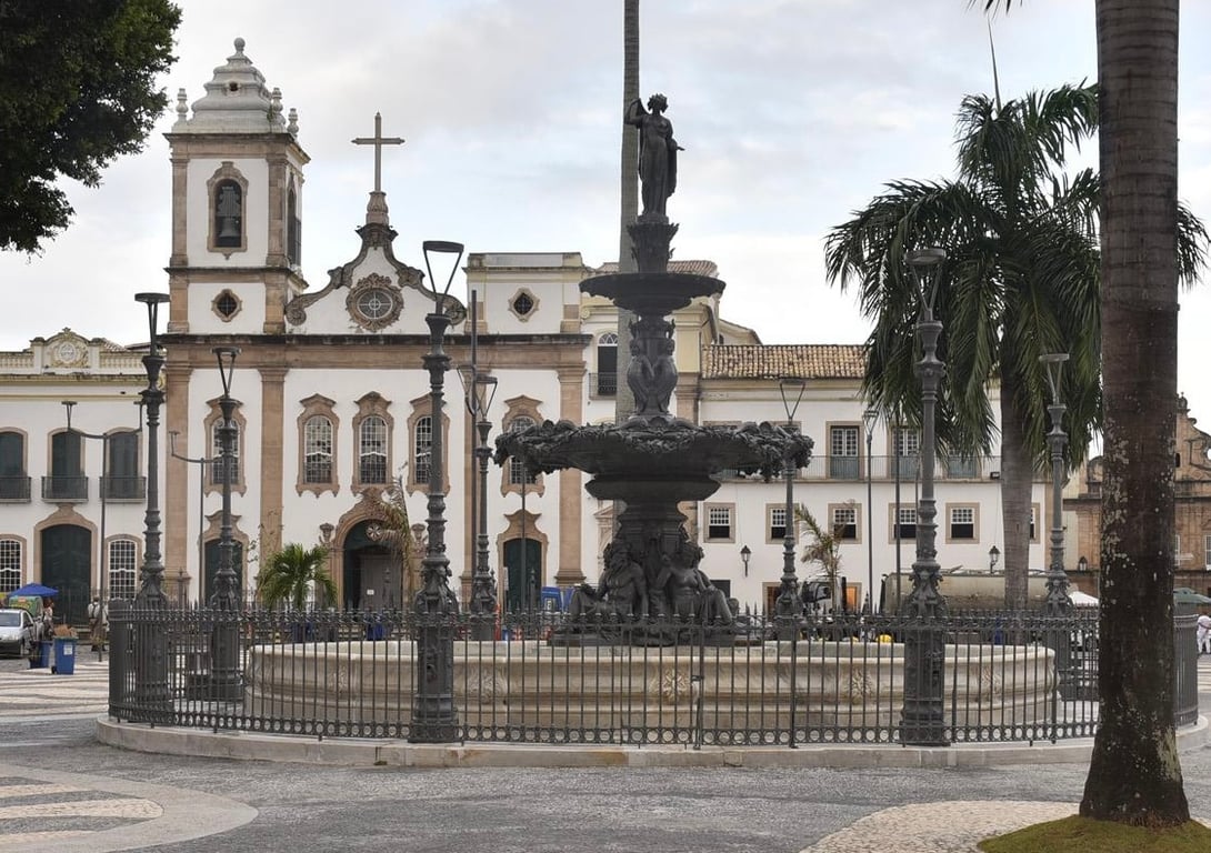 Fotografia atual do Chafariz do Terreiro de Jesus, em Salvador, publicada pelo portal Pelourinho Noite e Dia. A imagem mostra, em primeiro plano, um chafariz de ferro cercado por um gradil metálico. A estrutura possui dois níveis circulares e, no topo, uma escultura feminina sobre uma coluna central. Ao redor do chafariz, há postes de iluminação e palmeiras. Ao fundo, destaca-se a fachada da Catedral Basílica de Salvador, com sua torre, janelas e cruz no ponto mais alto, além de outros edifícios históricos que compõem o conjunto arquitetônico do Terreiro de Jesus. A fotografia registra um dos principais chafarizes públicos de Salvador, espaço que, durante o século XIX, era utilizado por aguadeiros e pela população para o abastecimento de água.