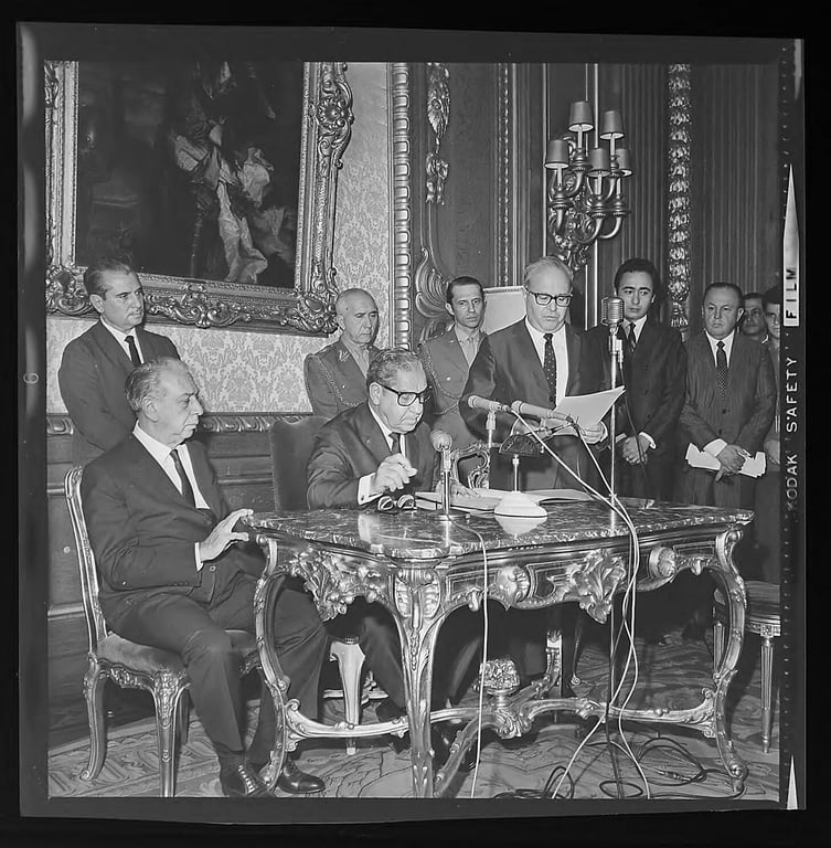 Fotografia em preto e branco registrada durante a cerimônia de instalação do Conselho de Defesa dos Direitos da Pessoa Humana (CDDPH), no Rio de Janeiro, em 1968. Em primeiro plano, ao centro da imagem, está sentado o então presidente do Brasil, o general Arthur da Costa e Silva, vestindo terno escuro e assinando um documento sobre uma mesa ornamentada de estilo clássico. À sua esquerda, também sentado, outro homem observa atentamente. Em pé, à direita de Costa e Silva, está o ministro da Justiça Luís Antônio Gama e Silva, lendo em voz alta um documento diante de diversos microfones. Atrás deles, há outros homens de terno e dois militares fardados, todos assistindo à cerimônia. O ambiente é luxuoso, com parede revestida por papel decorativo, molduras douradas e uma pintura clássica ao fundo. A cena revela o caráter formal e autoritário do evento.