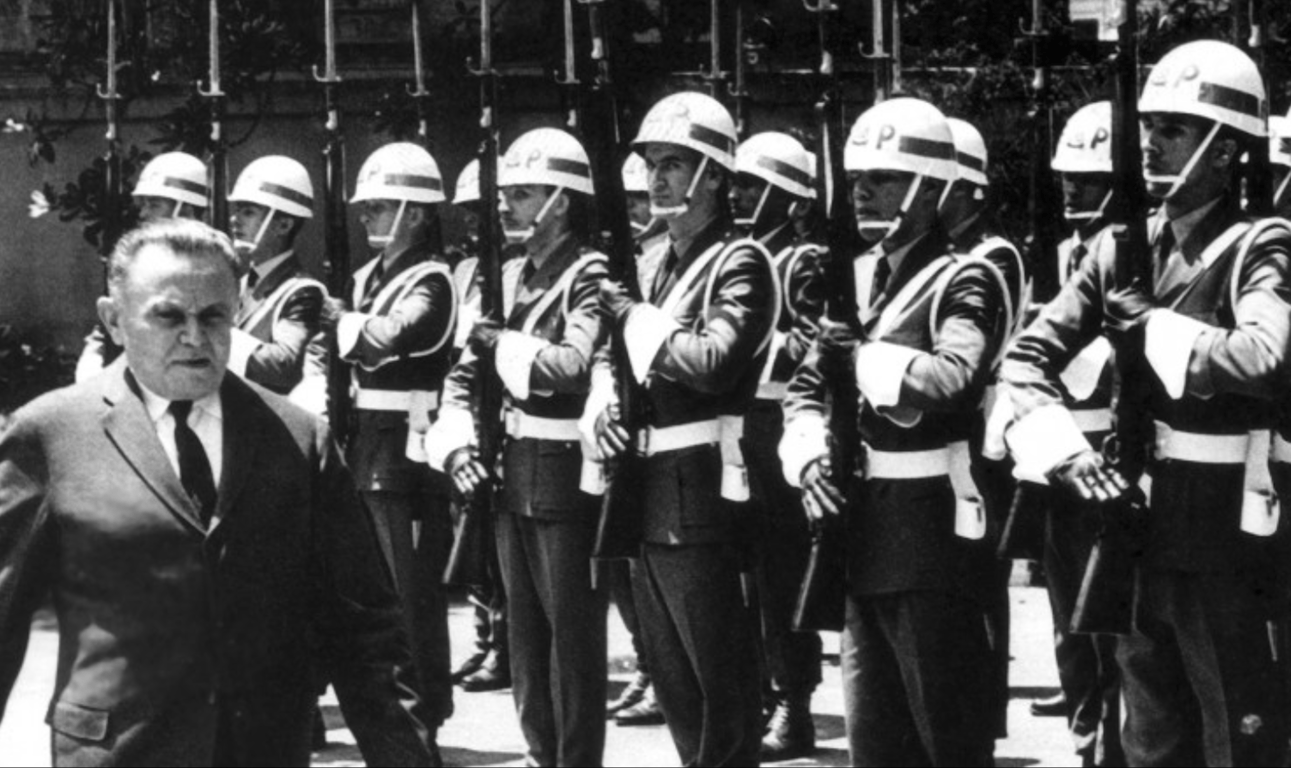 Fotografia em preto e branco mostra um homem de terno escuro caminhando à frente de uma fileira de soldados uniformizados. O homem aparenta ser uma figura de autoridade ou político, com expressão séria e olhar firme. Atrás dele, os soldados estão perfilados em posição de sentido, segurando fuzis com baionetas apontadas para cima. Eles vestem uniformes escuros com capacetes brancos que possuem a inscrição "PM" (Polícia Militar). Todos estão alinhados em formação rígida, transmitindo uma atmosfera de disciplina militar. A cena parece capturar um momento oficial ou cerimonial, possivelmente durante o período da ditadura militar no Brasil. Ao fundo, há árvores e uma construção parcialmente visível.