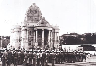 Fotografia em preto e branco, tirada no início do século XX, no Rio de Janeiro. Em primeiro plano, há uma formação de soldados uniformizados da Brigada Policial, alinhados em fileiras, de costas para a câmera. Os homens usam uniformes claros e capacetes arredondados, e alguns carregam armas. Ao fundo, destaca-se o imponente Palácio Monroe, edifício de arquitetura eclética com elementos neoclássicos e barrocos, incluindo colunas, esculturas e uma grande cúpula central.
