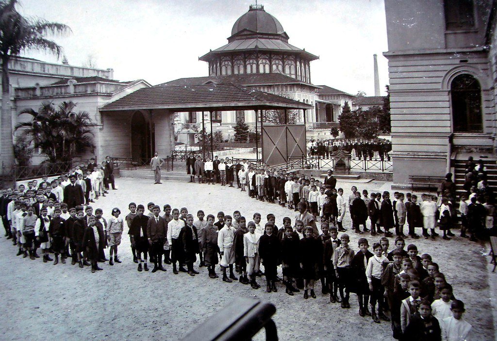 Fotografia em preto e branco de dezenas de meninos organizados em várias filas compridas no pátio de uma escola no final do século XIX. As crianças, de diferentes idades, estão vestidas com roupas sociais, como camisas, casacos, calças e alguns com boinas. Ao fundo, há um grande edifício com colunas e cúpula central, rodeado por construções menores, muros baixos, grades de ferro e vegetação. A cena mostra um momento de disciplina e organização escolar, onde a luz natural ilumina o ambiente externo, e a arquitetura ao fundo reforça o estilo monumental típico de instituições públicas da época.