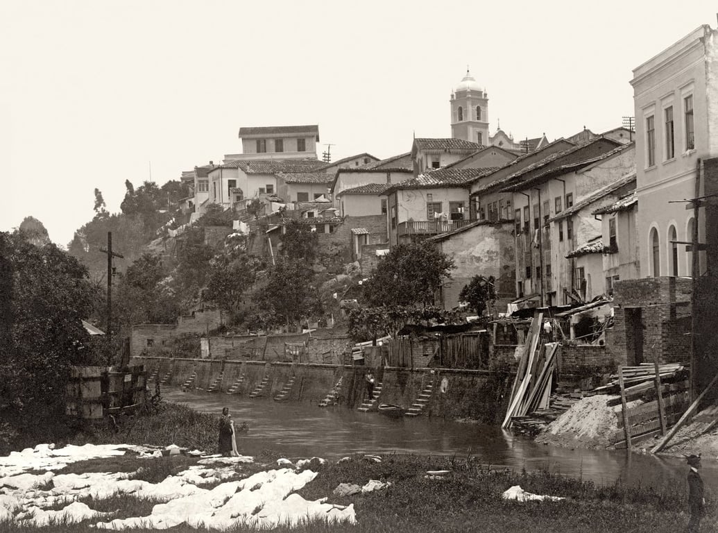 Fotografia em preto e branco do rio Tamanduateí, na cidade de São Paulo no início do século XX. À direita da imagem, há uma fileira de casas coloniais, algumas com fachadas deterioradas, construídas em desnível ao longo de um barranco à beira de um rio. No canto inferior esquerdo, uma mulher negra está em pé na margem do rio, com os pés dentro da água. Ela usa um vestido escuro e parece lavar roupas ou observá-las, pois há várias peças brancas estendidas no chão ao seu redor. Ao fundo, um conjunto de casas sobe a colina, formando um casario denso, com telhados de barro e construções de alvenaria. No topo da colina, destaca-se uma torre com cúpula, provavelmente pertencente a uma igreja.