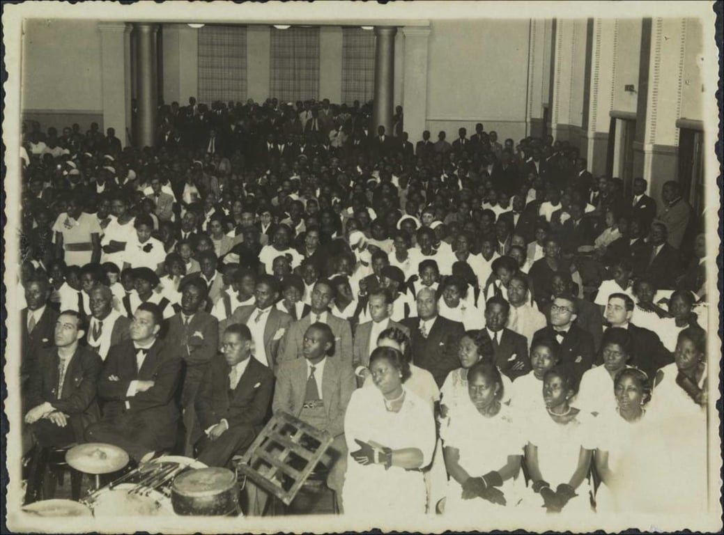 Fotografia em preto e branco registra uma multidão de pessoas negras reunidas em um salão amplo, com colunas altas e janelas ao fundo. A imagem mostra parte da assistência presente na comemoração do aniversário de fundação da Frente Negra Brasileira, realizado em 16 de setembro de 1935, em São Paulo (SP). Na composição, homens e mulheres aparecem em trajes formais — ternos, vestidos claros, luvas e adornos no cabelo — refletindo a seriedade e solenidade do evento. À frente, instrumentos de percussão como tambores e pandeiros sugerem a presença de música ou apresentações culturais. O salão está completamente lotado, com fileiras organizadas e grande concentração de pessoas também em pé ao fundo. A imagem expressa o protagonismo e a articulação política da população negra durante o período, sendo um marco da atuação da Frente Negra Brasileira, uma das mais importantes organizações de luta por direitos civis e sociais dos negros no Brasil da década de 1930.