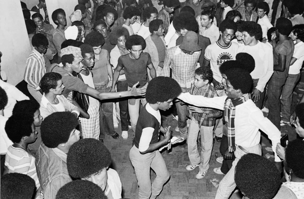 Fotografia em preto e branco de um baile black no Rio de Janeiro, provavelmente dos anos 1970. A imagem mostra dezenas de jovens negros dançando em uma pista de dança, vestindo roupas características da época. A atmosfera é de celebração e resistência cultural durante o período da ditadura militar.