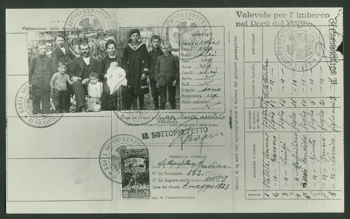 Fotografia de um passaporte italiano de 1923, acervo do Museu da Imigração de São Paulo. O documento, em preto e branco, contém escrita e impressa em italiano, com selos e carimbos oficiais espalhados pela página. No topo direito, há uma fotografia colada de um grupo de imigrantes italianos, entre homens, mulheres e crianças, todos vestidos com roupas de época e postados em posição formal para o registro. O fundo da foto exibe uma paisagem ao ar livre, sugerindo um ambiente rural ou periférico. O passaporte apresenta marcas de envelhecimento, como dobras e leve desgaste nas bordas.
