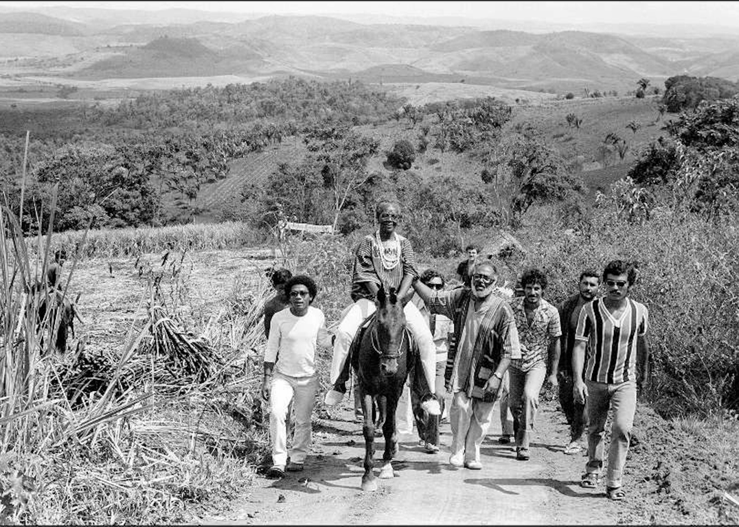 Fotografia em preto e branco retrata uma cena histórica no caminho do Quilombo dos Palmares, na Serra da Barriga. Ao centro, Mãe Hilda de Jitolu, ialorixá do Ilê Axé Jitolu, aparece montada em um cavalo, com expressão serena e vestida com bata estampada e colares rituais. Ela é ladeada por duas figuras importantes: à sua esquerda, Passarinho, e à sua direita, Abdias Nascimento, ambos caminhando próximos e sorridentes. Outros homens acompanham o grupo, andando pela estrada de terra que atravessa um cenário de roçados e mata. Ao fundo, a paisagem revela morros, vales e vegetação densa, compondo um ambiente que remete à memória da resistência quilombola.