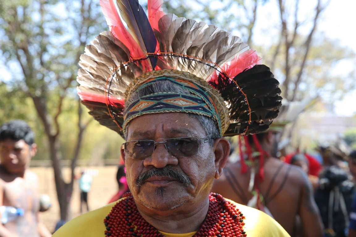A fotografia mostra um homem indígena mais velho, vestindo um cocar de penas e colares tradicionais, que posa para a câmera com uma expressão séria. A composição é focada no homem, com uma proximidade que preenche a maior parte do quadro. Ele está centrado, com o fundo desfocado, o que direciona a atenção para ele. Pessoas quilombolas podem ser vistas no fundo, um pouco desfocadas, indicando um evento.