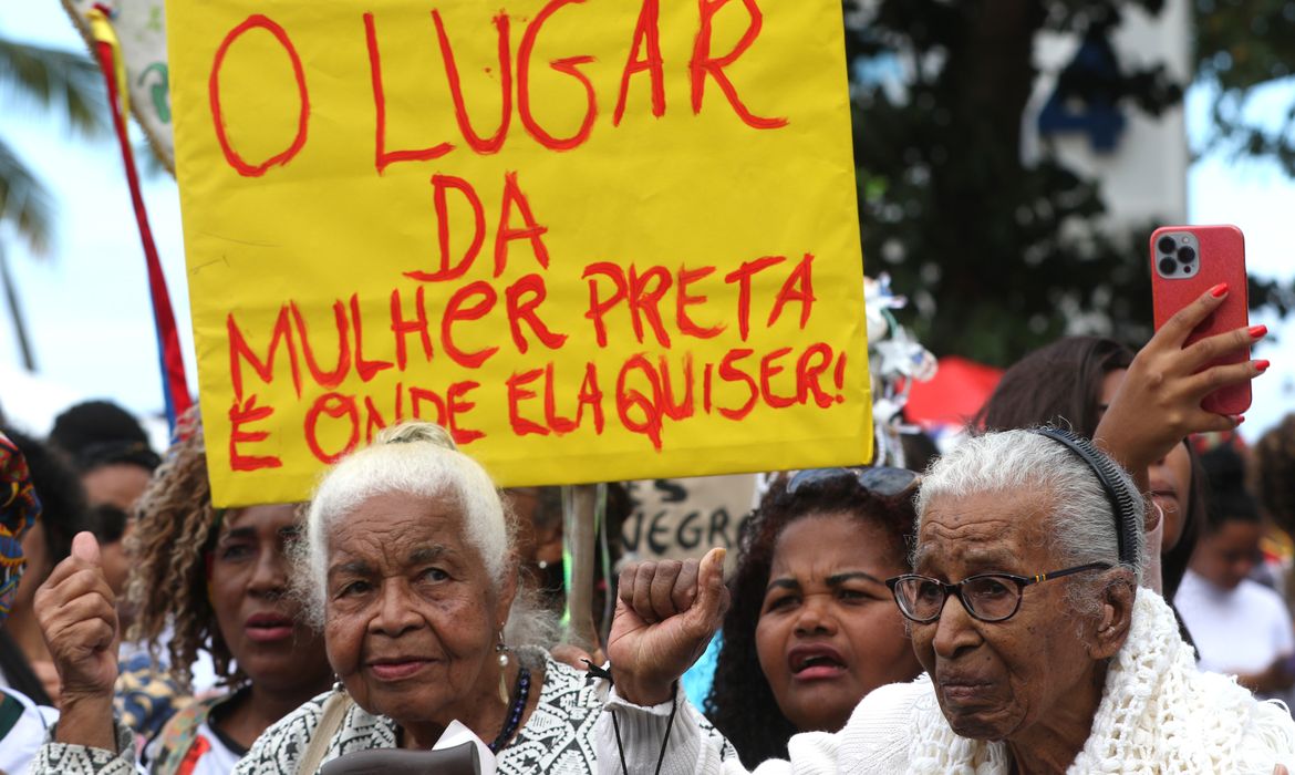 A fotografia mostra um grupo de mulheres em protesto, com foco em um cartaz amarelo que destaca a frase 'O lugar da mulher preta é onde ela quiser!'. A ação parece estar em andamento, com as pessoas parecendo estar em movimento ou atentas aos arredores. O enquadramento é em close-up, privilegiando as participantes da manifestação e o cartaz, com outras pessoas ao fundo. A interação entre as pessoas e o cartaz sugerem um ato de protesto político.