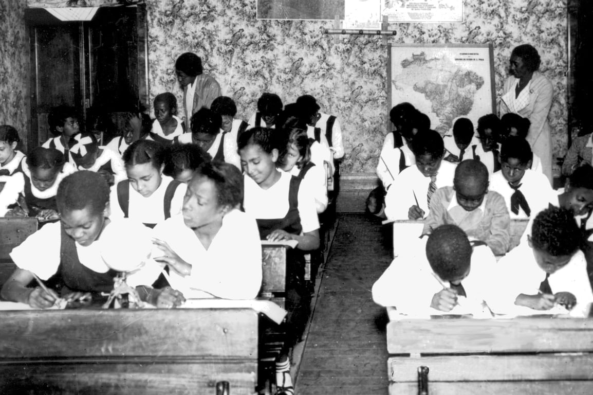 Fotografia em preto e branco de uma sala de aula de uma escola da Frente Negra Brasileira cheia de crianças negras, na primeira metade do século XX. As crianças estão sentadas em carteiras de madeira alinhadas em filas, escrevendo em cadernos, vestindo uniformes escolares (camisa branca e avental ou gravata). Duas professoras negras estão em pé na sala, observando e auxiliando os alunos. Ao fundo, é possível ver um mapa do Brasil pendurado na parede e papel de parede estampado com flores. A imagem transmite concentração, aprendizado e organização, refletindo um ambiente educacional estruturado.