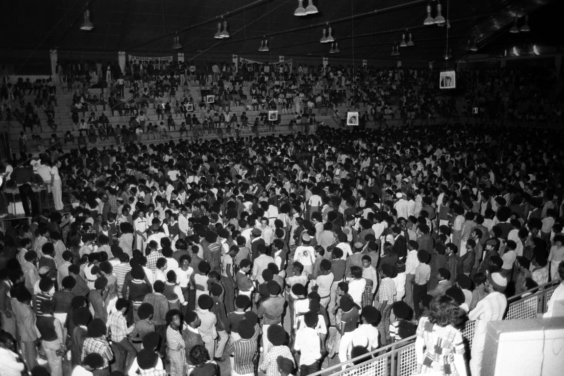 Fotografia em preto e branco mostra um grande ginásio lotado de pessoas durante um baile black carioca nos anos 1970. O espaço está completamente tomado por uma multidão majoritariamente composta por jovens negros e negras, muitos deles com cabelos no estilo afro. No centro e nas arquibancadas, as pessoas se aglomeram, algumas de pé e outras sentadas, criando uma atmosfera de energia e coletividade. Pendurados no teto, vêem-se quadros com rostos de personalidades negras, símbolos de referência e identidade. A imagem transmite força, resistência cultural e celebração da negritude.