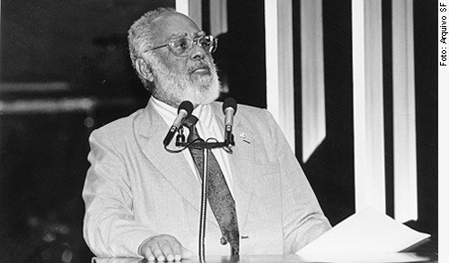 Fotografia em preto e branco de um homem negro idoso, de barba e cabelo grisalhos, falando ao microfone. Ele veste terno claro, camisa branca e gravata escura, e segura folhas de papel com a mão esquerda enquanto apoia a direita sobre o púlpito. Ao fundo, há barras verticais claras em contraste com o fundo escuro. A imagem foi registrada durante um discurso no Senado Federal entre 1997 e 1999. Trata-se de Abdias Nascimento, importante político, artista e ativista do movimento negro no Brasil. À direita da imagem, lê-se 'Foto: Arquivo S.F.' em letras verticais.