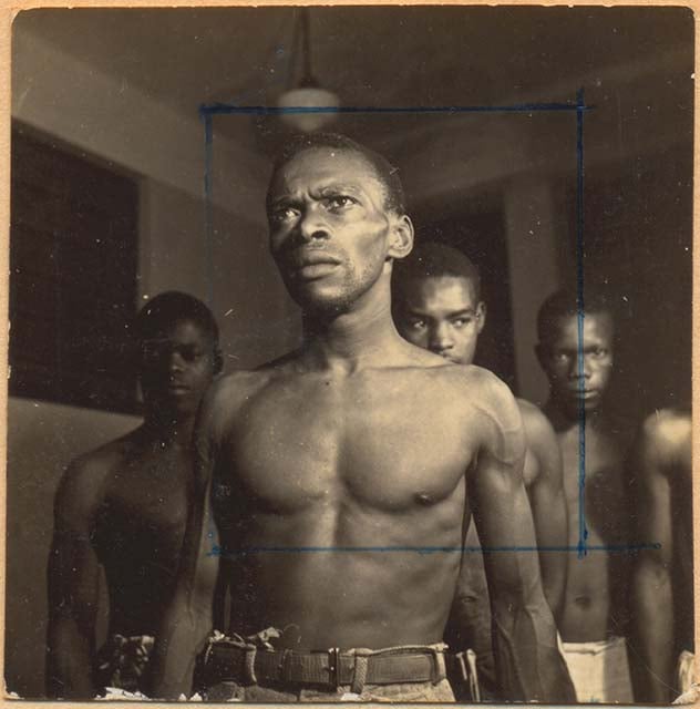 Fotografia em preto e branco retrata cinco homens negros, de tronco nu, posicionados em ambiente interno com iluminação central. O homem em primeiro plano está à frente dos demais, com olhar firme e expressão séria, evidenciando sua força e postura determinada. Ele veste calça com cinto e está centralizado dentro de um enquadramento retangular traçado à caneta azul diretamente sobre a imagem. Ao fundo, os outros quatro homens observam com semblantes concentrados. A imagem sugere um registro oficial ou científico, possivelmente ligado a estudos de eugenia ou antropometria do século XX.