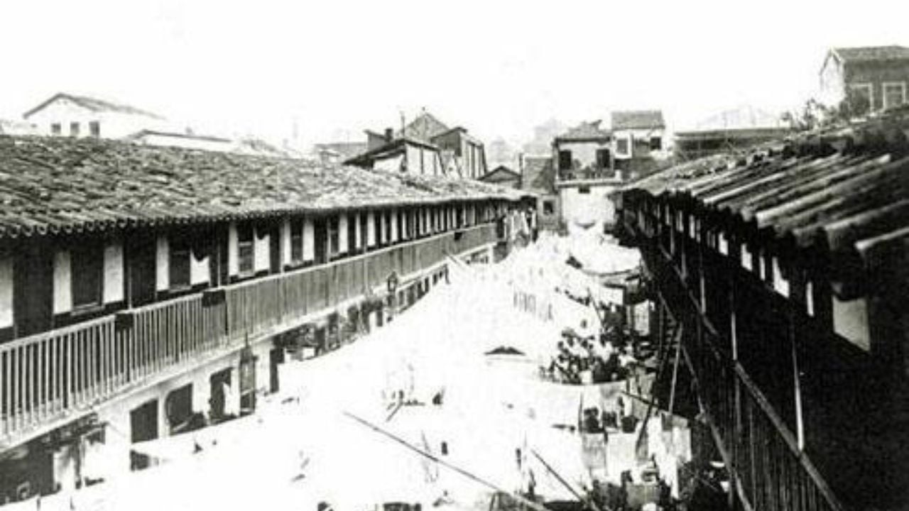 Fotografia em preto e branco intitulada 'O Cortiço Cabeça de Porco', da Biblioteca Nacional. A imagem retrata um cortiço do Rio de Janeiro na década de 1880, com longos corredores de madeira ladeados por pequenas moradias. No centro, um espaço aberto com roupas estendidas e pessoas circulando. Ao fundo, edificações da cidade.