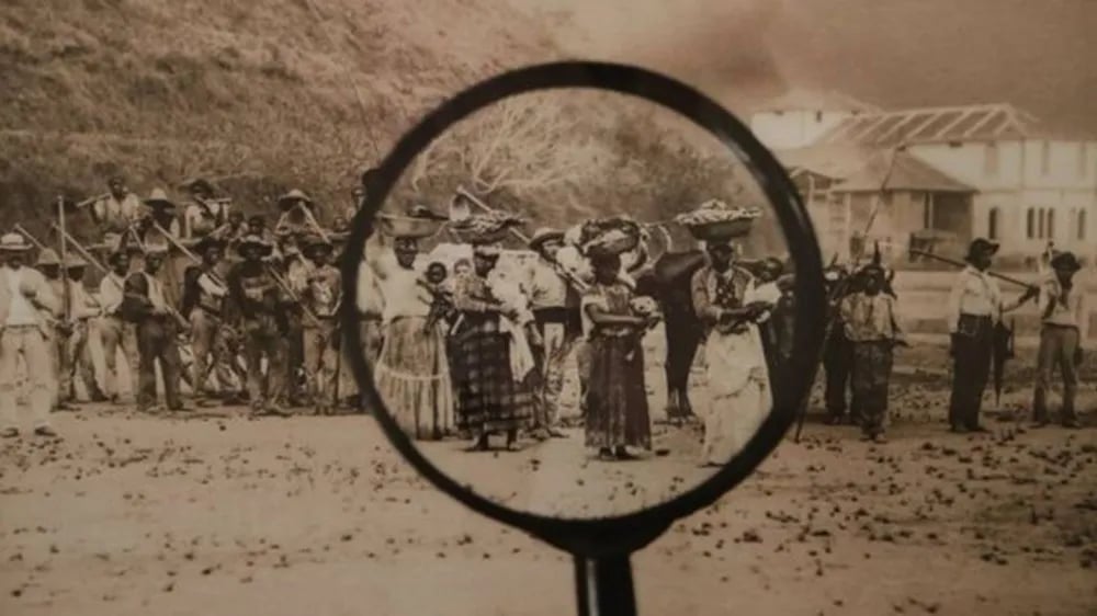 Fotografia histórica “Ganhadores”, atribuída a Albert Frisch, Marc Ferrez e/ou Paul G. L. G. Lofgren, reproduzida pela BBC. A cena mostra um grupo de homens, mulheres e crianças negras reunidos em uma área aberta. Em primeiro plano, uma lupa amplia parte da fotografia, destacando mulheres negras que carregam cestos e trouxas sobre a cabeça, acompanhadas por crianças e outras pessoas ao redor. À esquerda, um grupo de homens segura ferramentas de trabalho apoiadas nos ombros. À direita, aparecem outros trabalhadores reunidos próximos a construções. Ao fundo, há árvores e edifícios. A fotografia registra homens e mulheres conhecidos como ganhadores, trabalhadores que realizavam o transporte de mercadorias, alimentos e outros produtos nas cidades brasileiras durante o século XIX.