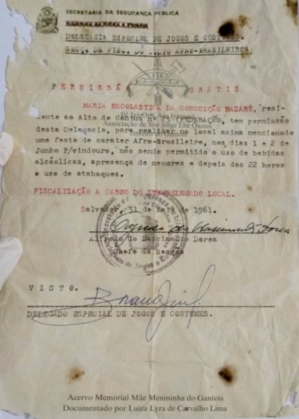 Documento intitulado Permissão para Realização de Festa de Caráter Afro-Brasileiro, pertencente ao acervo do Memorial Mãe Menininha do Gantois. O documento, datado de 31 de maio de 1961, é datilografado em papel de tom amarelado, com marcas de envelhecimento, dobras, manchas e pequenos rasgos nas bordas. Na parte superior, aparecem o brasão do Estado da Bahia e o cabeçalho da Secretaria da Segurança Pública, com a identificação da Delegacia Especial de Jogos e Costumes. No centro, o texto concede autorização para Maria Escolástica da Conceição Nazaré realizar uma festa de caráter afro-brasileiro nos dias 1º e 2 de junho, estabelecendo condições como a proibição do uso de bebidas alcoólicas, da presença de menores após as 22 horas e do uso de atabaques. Na parte inferior, há carimbos oficiais, assinaturas manuscritas e a identificação do acervo do Memorial Mãe Menininha do Gantois, com documentação de Luiza Lyra de Carvalho Lima.
