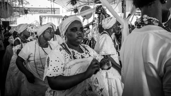 Fotografia em preto e branco de um culto de religiões afro-brasileiras, de Tiago Celestino, publicada no Unsplash. A imagem mostra, em primeiro plano, uma mulher negra vestida com roupa branca rendada, pano da costa e torço na cabeça. Ela segura um pequeno objeto com as duas mãos e olha para a frente. Ao seu redor, outras mulheres negras também usam vestimentas brancas tradicionais, torços e colares de contas, participando da cerimônia. À direita, aparece parcialmente uma pessoa de costas, usando roupa branca e fios de contas no pescoço. Ao fundo, observam-se mais participantes e elementos do espaço religioso, registrados durante um momento de celebração de uma religião de matriz africana.