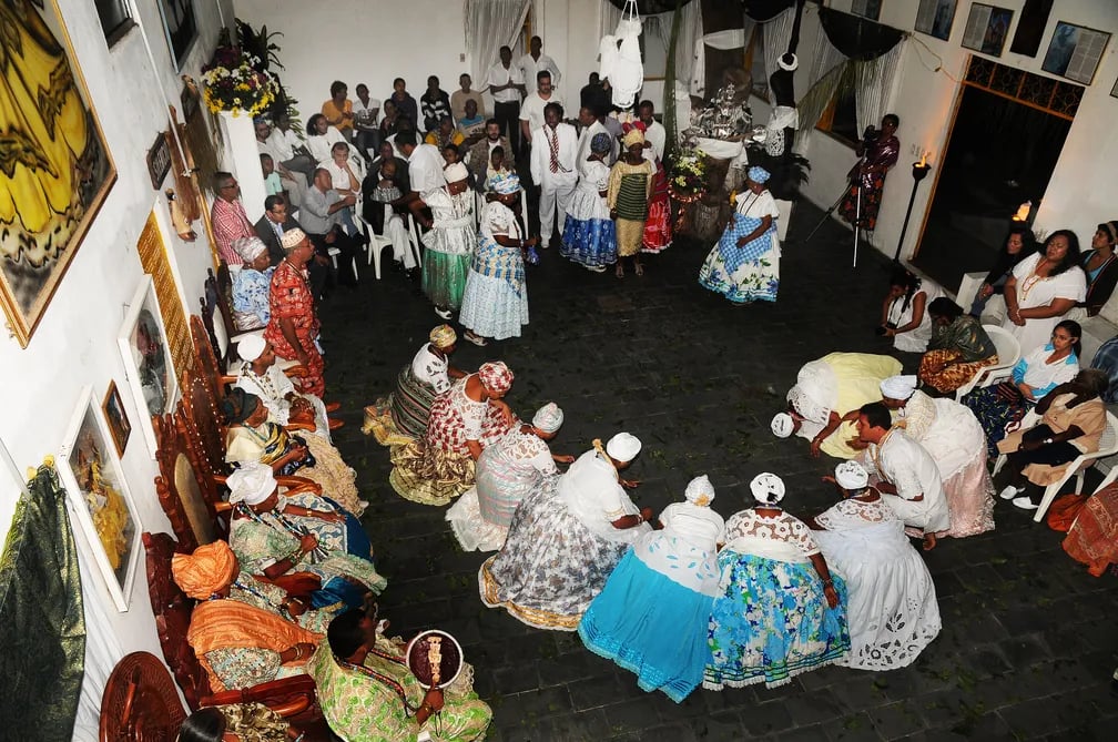 Fotografia do Terreiro Raiz de Ayrá, na cidade de Cachoeira, Bahia, de Lázaro Menezes, produzida para a Secretaria de Cultura da Bahia. A imagem mostra o interior de um terreiro de candomblé durante uma cerimônia religiosa. No centro do salão, homens e mulheres, em sua maioria pessoas negras, formam uma grande roda. Muitos vestem roupas brancas e trajes tradicionais do candomblé, enquanto algumas mulheres usam saias rodadas coloridas, panos da costa e torsos na cabeça. Nas laterais, outras pessoas permanecem sentadas em cadeiras ou bancos, acompanhando a celebração. Ao fundo, há um altar decorado com flores, tecidos brancos e objetos litúrgicos. As paredes são brancas e exibem quadros e elementos decorativos. A fotografia registra um momento de celebração e prática da religiosidade de matriz africana em um dos tradicionais terreiros da cidade de Cachoeira.